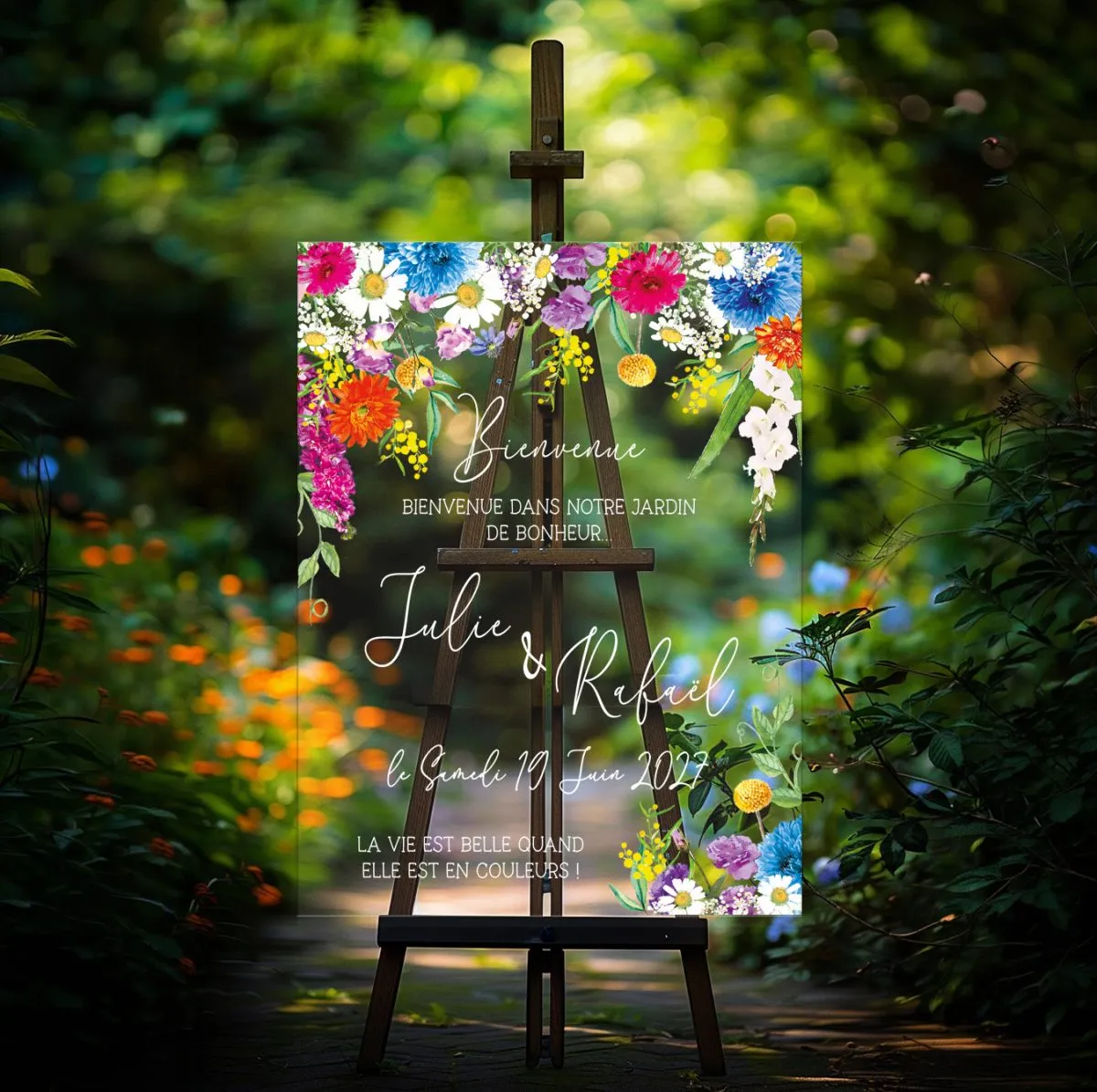 Panneau de bienvenue mariage en plexiglas transparent, illustration fleurs sauvages multicolores gerberas orange bleuets et marguerites aquarelle, sur chevalet bois foncé dans une allée de jardin sombre et verdoyante, personnalisé aux prénoms des mariés